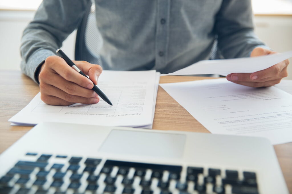 Pessoa analisando documentos, com uma caneta na mão e em frente a um notebook, representando onde fazer tradução juramentada.
