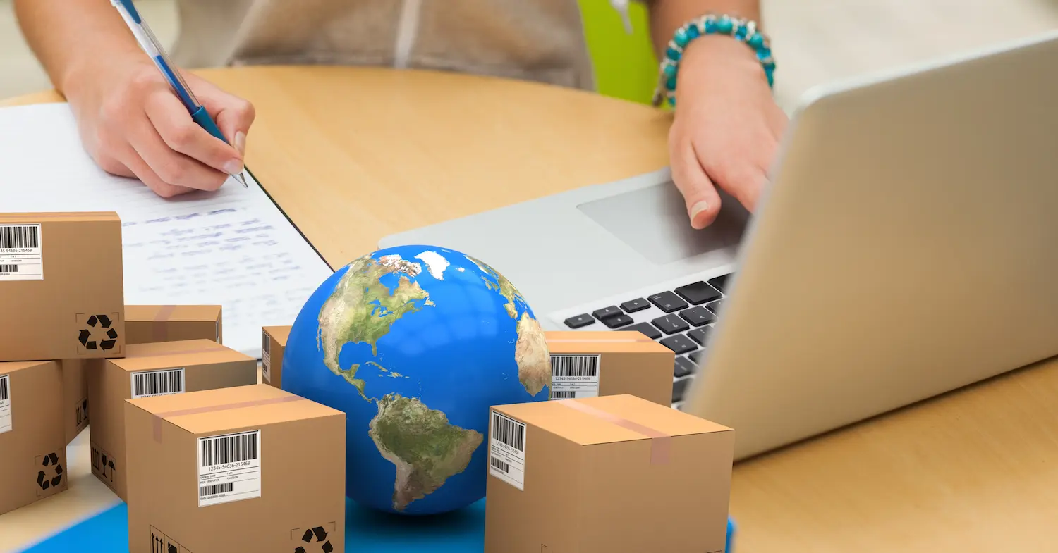Pessoa fazendo anotações de como exportar produtos, com notebook, miniaturas de caixas e do globo sobre a mesa.