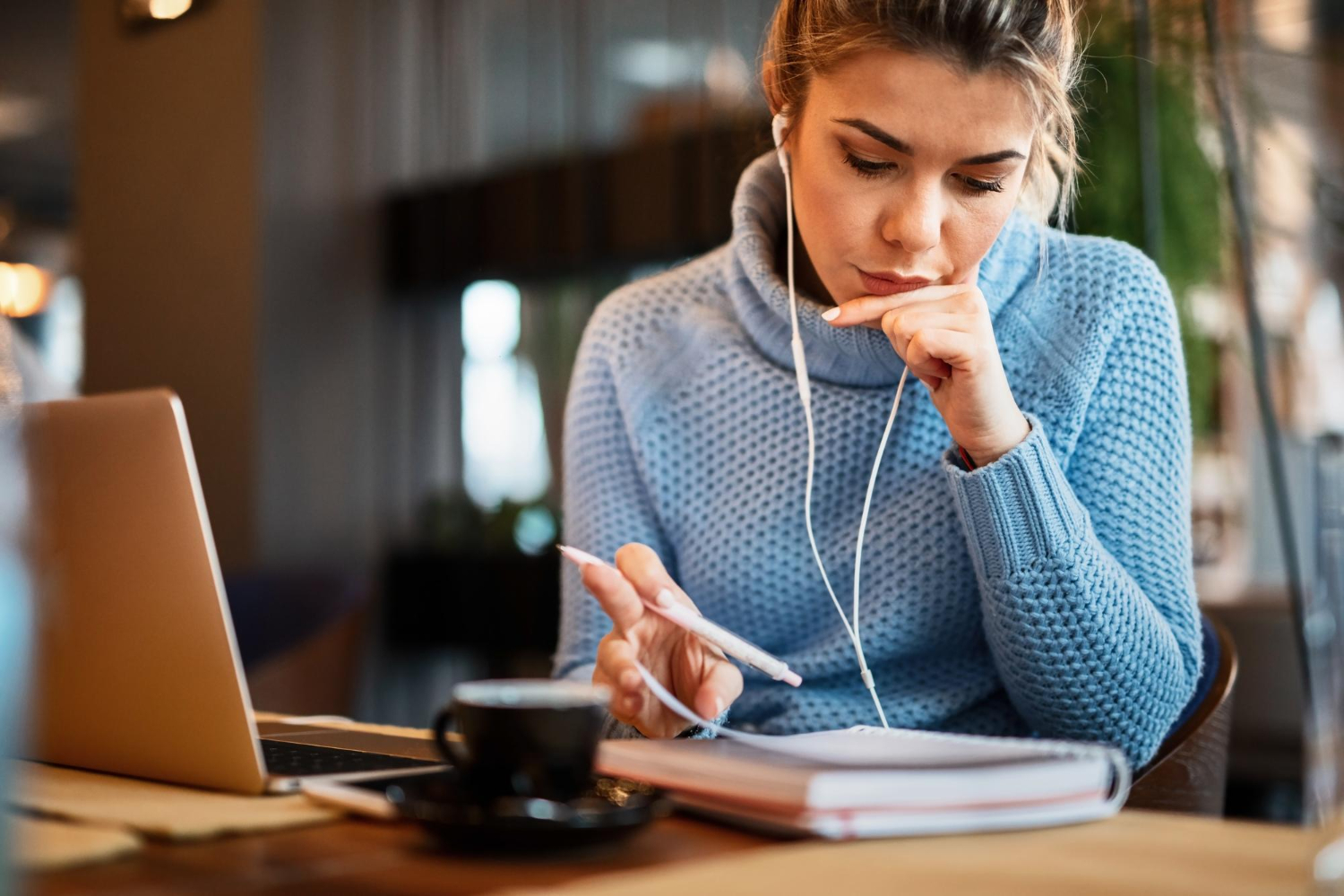 Mulher branca de blusa azul com fones de ouvido sentada olhando um caderno e trabalhando com a transcrição fonética.
